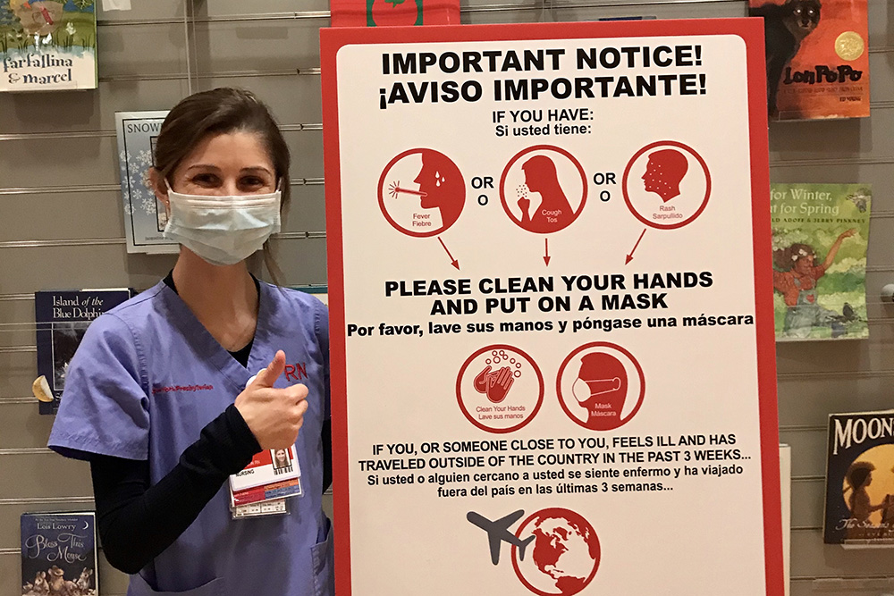 UVA Nursing grad Lauren Dickinson (BSN `19) standing next to poster warning people to wash hands and put on a mask to prevent spreading the 2020 COVID-19 pandemic