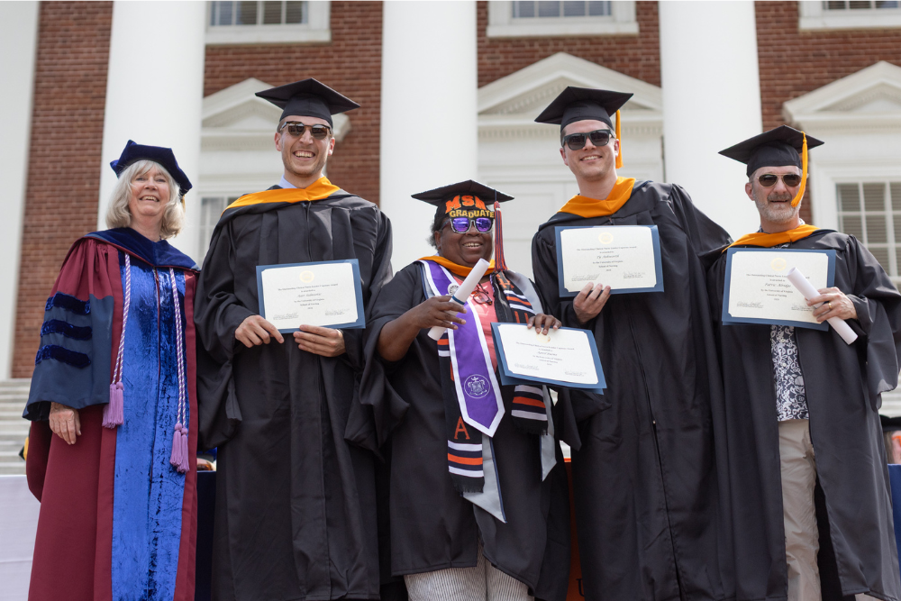 Four nursing student graduates standing in cap and gown with their diplomas next to the dean on the UVA Rotunda steps