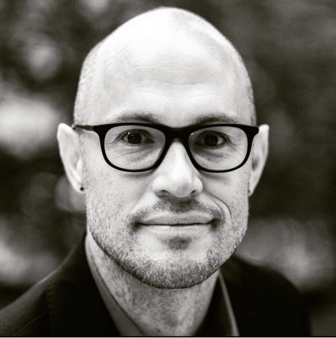 A black and white image of a bald guy wearing glasses smirking into the camera