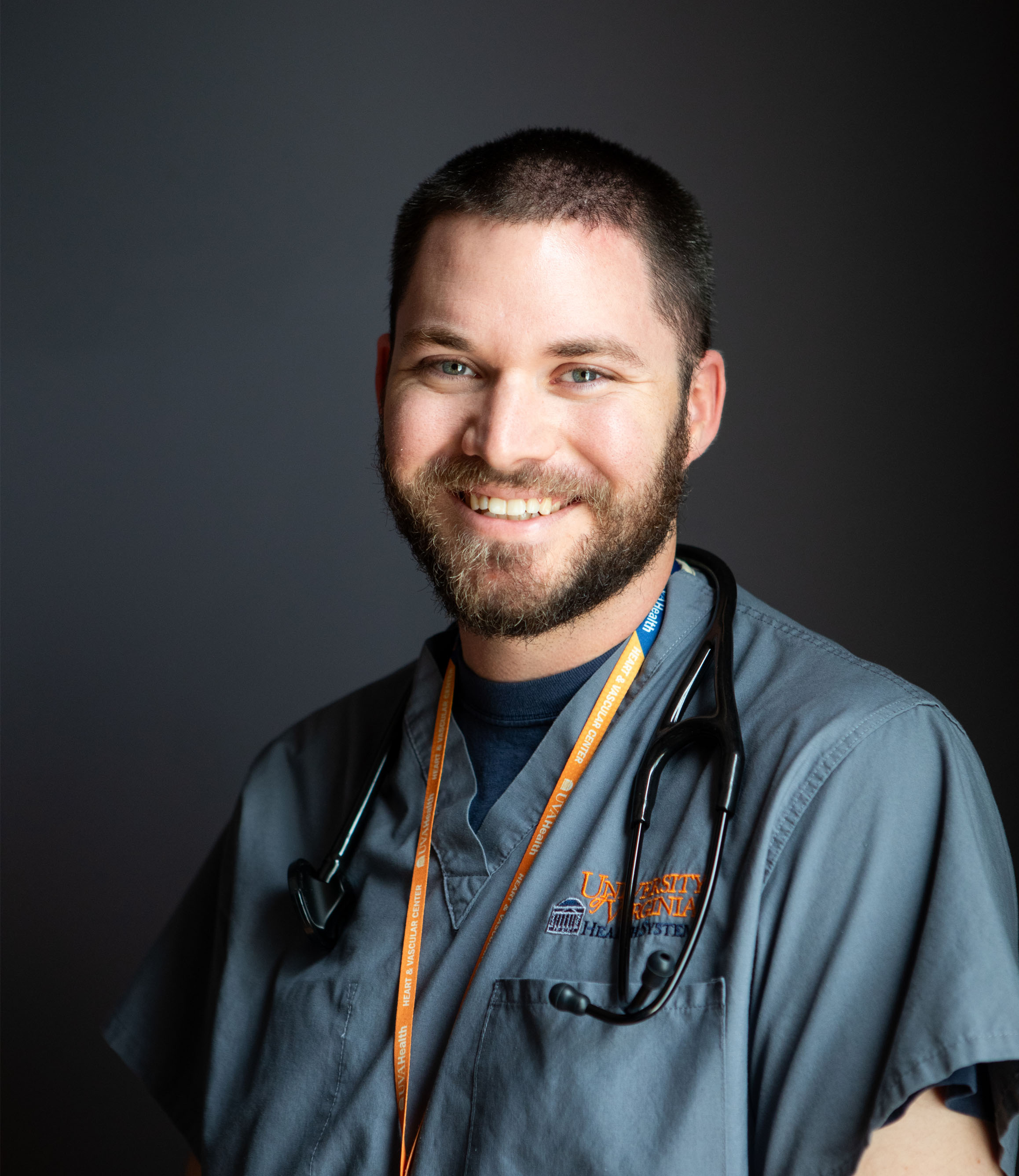 A guy smiling in scrubs with short brown hair wearing a stethoscope